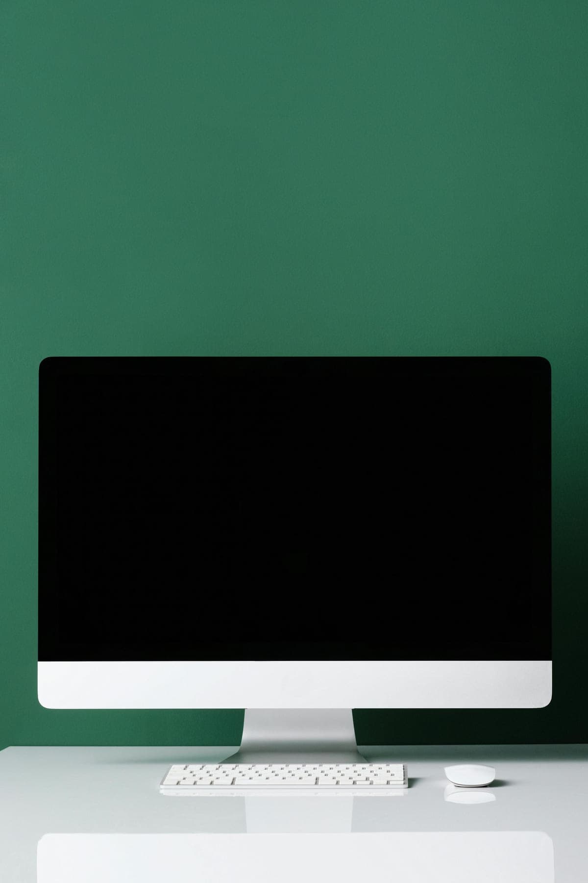 A clean desktop monitor against a forest-green wall, screen black, keyboard ready